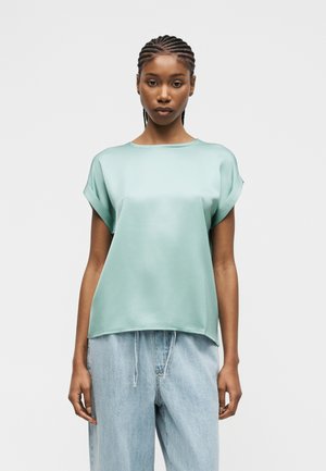 Kvinde med fletninger iført en løs, mintgrøn satinbluse med korte ærmer og lyseblå højtaljede jeans mod en ensfarvet hvid baggrund.