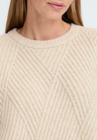 Beige gestrickter Pullover mit strukturiertem Rippmuster, ausgestattet mit einem runden Ausschnitt und diagonalen Stichmustern über den Stoff.
