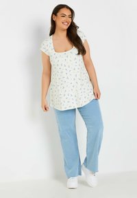 Túnica floral de manga corta, fondo crema claro, con un patrón floral en azul. Combinada con unos jeans acampanados azul claro y zapatillas blancas.