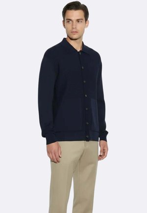 Uomo che indossa un cardigan blu scuro abbottonato con tasche e pantaloni beige, in piedi e che guarda di lato su uno sfondo uniforme.