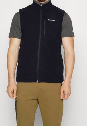 Man draagt een zwarte Columbia fleece bodywarmer met rits aan de voorkant over een grijs T-shirt met korte mouwen en bruine broek met zijvakrits.