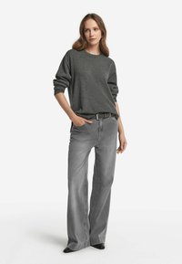 Grauer Oversize-Pullover mit umgeschlagenen Bündchen, kombiniert mit lässigen grauen Jeans und schwarzen Schuhen, vor einem einfachen weißen Hintergrund.