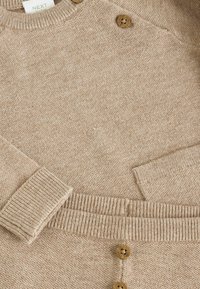 Vaahto-punottu beige kardigan, jossa on raidoitukset hihansuissa ja kaula-aukossa. Edessä neljä ruskeaa nappia, joissa on teksturoitu kangaskudos.