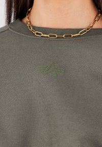 Olíva zöld pulóver bordázott textúrával és kerek nyakkivágással, a mellrészén finom, azonos színű, hímzett logóval. Aranylánc nyaklánc.