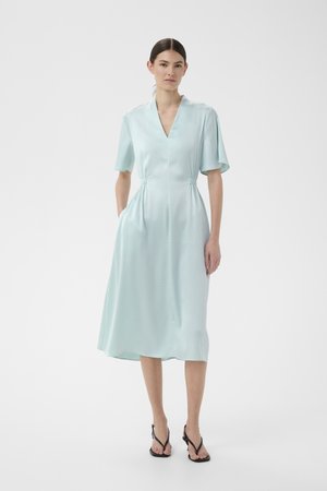 Ženska nosi svetlo modro midi obleko s kratkimi rokavi, V-izrezom in žepi, kombinirano s črnimi sandali z naramnicami na belem ozadju.