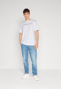 Šviesiai pilkas medvilninis marškinėlis su juodu "TOMMY JEANS" logotipu, derinamas su šviesiai mėlynomis džinsinėmis kelnėmis ir baltais sportbačiais ant vientiso fono.