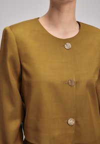 Senapsgul jacka med rund halsringning, tre dekorativa guldfärgade knappar och en texturerad tygfinish; långa, något puffiga ärmar.
