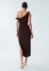 Donna con capelli raccolti in uno chignon, che indossa un vestito aderente di colore marrone scuro a una spalla, con una spacco laterale e sandali dorati con tacco alto, in piedi davanti a uno sfondo neutro.