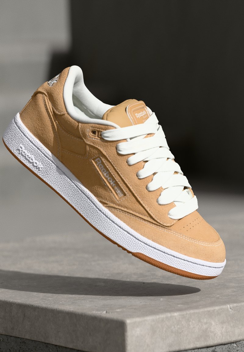Sneaker Reebok in suede color tan con lacci e suola bianchi, bilanciata su un ledge di pietra grigia con uno sfondo scuro sfocato.