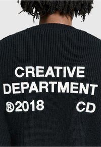 Black knitted cardigan featuring white embroidered text that reads “CREATIVE DEPARTMENT,” “®2018,” and “CD.” Text has a textured appearance.