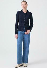 Marinblå räfflad långärmad cardigan med krage, med knappstängning fram. Kombinerad med ljusblå vida jeans och beige skor.