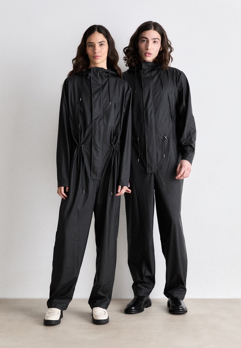 Deux personnes portent des vêtements d'extérieur noirs assortis, avec des détails à cordon de serrage. Le tissu semble élégant, avec des poches et des coupes confortables et amples.