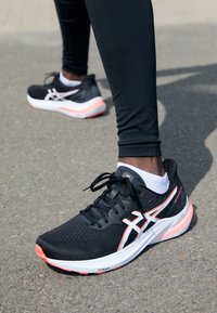 Zapatillas deportivas negras con acentos en blanco y rosa, parte superior texturizada, diseño de cordones y suelas acolchadas, ubicadas sobre una superficie exterior gris.