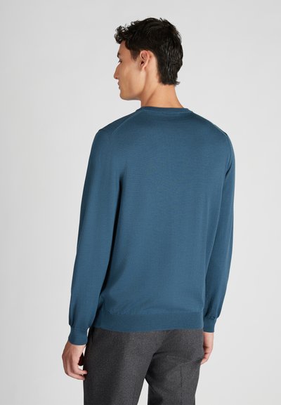 Pullover blu a maniche lunghe in maglia strutturata, con scollatura rotonda e polsini a costine. Indossato con pantaloni grigi.