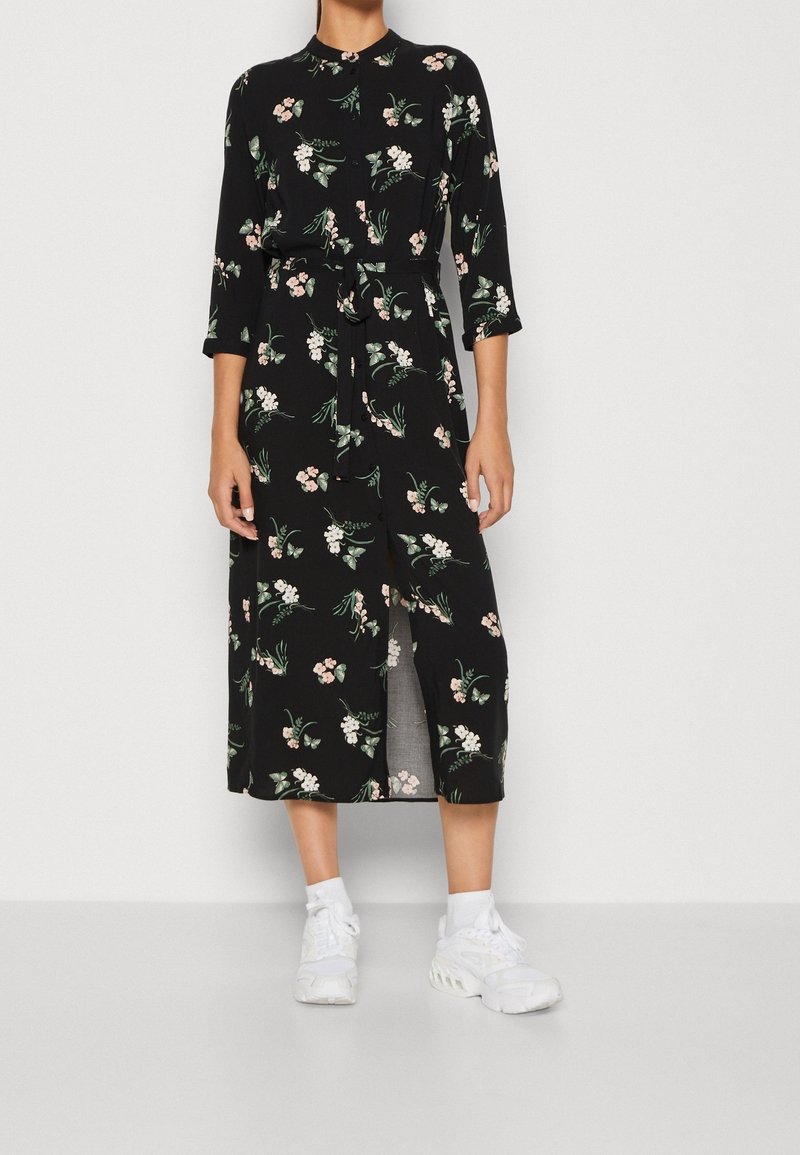 Personne portant une robe midi noire avec des motifs floraux roses et blancs, une fente devant, manches trois-quarts, chaussettes blanches et baskets blanches.