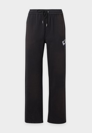Jeune homme portant une casquette noire, un sweat à capuche zippé noir avec des patchs, un t-shirt blanc, un pantalon noir avec un texte blanc, et des baskets noir et blanc.