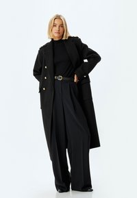 Abrigo largo de lana negra con botones dorados, usado sobre una blusa negra y pantalones de pierna ancha negros de cintura alta. Cinturón simple y textura lisa.