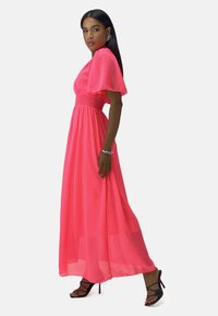 Robe maxi de couleur rose vif, en tissu léger. Dotée d'une taille smockée et de manches volantes. Associée à des sandales noires à talons hauts.