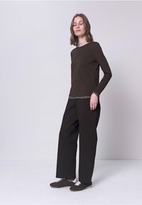 Vrouw staand met gesloten ogen, dragend een zwart top met lange mouwen en patroon op de manchetten, zwarte brede broekspijpen en zwarte sneakers.