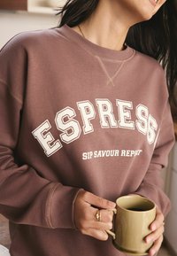 Brauner Pullover mit weißen Buchstaben, auf denen "ESPRESS" und "SIP SAVOUR REPEAT" steht. Glatte Textur, lässiger Schnitt und gerippte Bündchen. Person, die eine grüne Tasse hält.