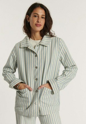 Kobieta ubrana w jasnobłękitną i beżową marynarkę w pionowe paski z guzikami oraz pasujące spodnie, ręce w kieszeniach, z krótkimi ciemnymi włosami.