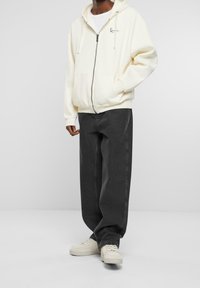Sudadera crema con cremallera y cordones, que presenta un logo en el frente. Combinada con pantalones vaqueros holgados de color gris oscuro y zapatillas de color claro.