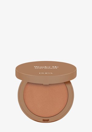 Compact bronzer met een zijdezachte, romige textuur. Heeft een glanzend, bruin poeder en een ronde beige verpakking met het label "Wonder Me" duidelijk zichtbaar.