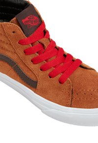 Baskets montantes en suede marron avec lacets rouges et une bande latérale marron. Elles sont dotées d'un col rembourré et d'une semelle en caoutchouc blanc avec une texture antidérapante.