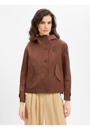 Frau trägt eine braune, mit Knöpfen versehene Jacke mit Klappentaschen und beigefarbene, weit geschnittene Hosen vor einem einfarbigen weißen Hintergrund.