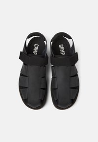 Black sandals made of soft material with adjustable straps. Features a closed toe and open heel design, highlighting a flexible and breathable construction.