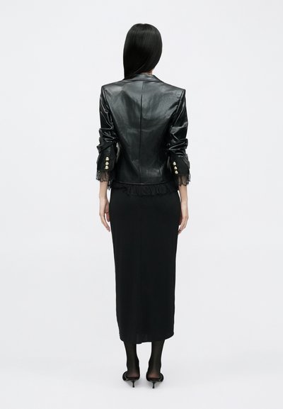 Fekete bőrkabát rojtokkal, arany gombokkal és testhez simuló szabással, hosszú fekete szoknyával és áttetsző fekete harisnyával párosítva.