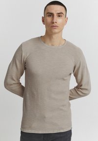 Beige long-sleeve t-shirt with a round neck, textured fabric, and subtle horizontal pattern. Styled with dark jeans.