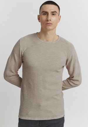 Beige long-sleeve t-shirt with a round neck, textured fabric, and subtle horizontal pattern. Styled with dark jeans.