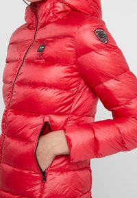 Rode gewatteerde jas met glanzende afwerking, horizontale stiksels, met ritssluiting afsluitbare zijzakken en een logopatch op de schouder.