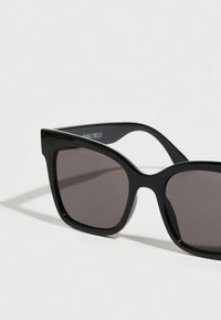 Μαύρα υπερμεγέθη γυαλιά ηλίου με γυαλιστερή επιφάνεια, με ορθογώνια πλαίσια και σκουρόχρωμους φακούς, με την επιγραφή "ANNA FIELD" στο εσωτερικό του βραχίονα.