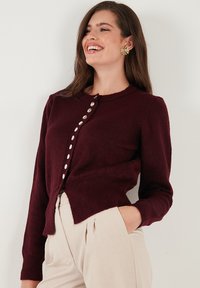 Cardigan en tricot bordeaux avec un col rond, des boutons en fausse perle sur le devant, des poignets côtelés et une coupe légèrement courte.