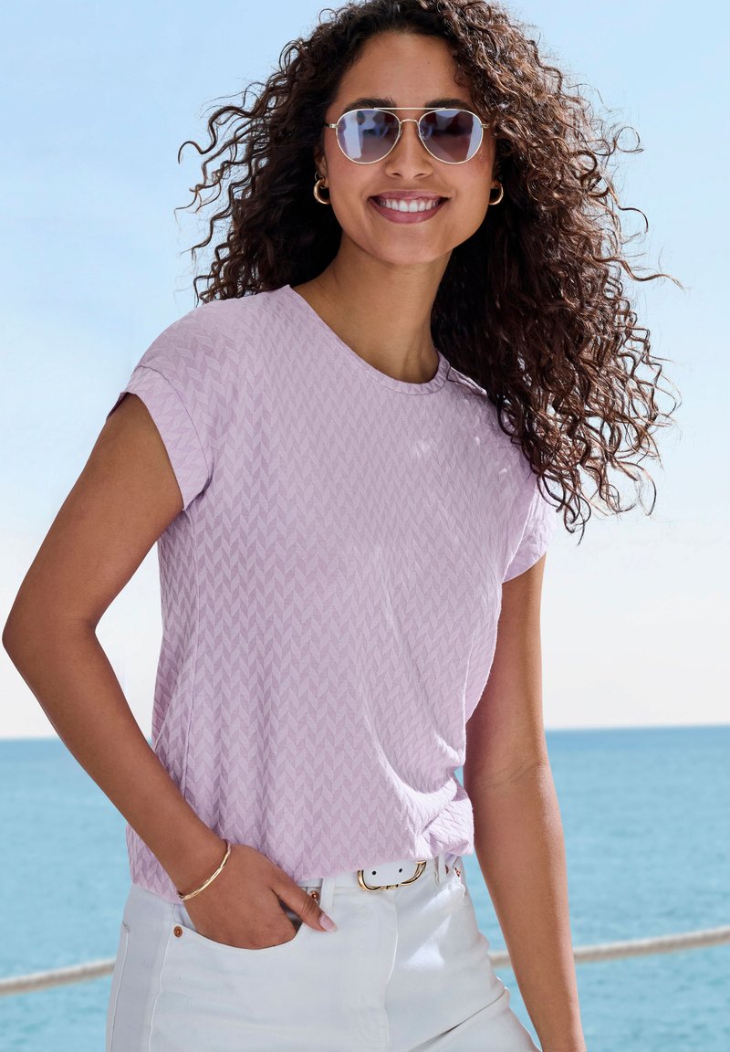 T-shirt violet clair avec un motif en zigzag texturé, col rond, manches courtes, associé à un jean blanc et une ceinture rehaussée d'or.