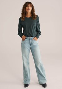 Sötétzöld, hosszú ujjú felső, vállain összegyűrt részletekkel, világoskék, bő szárú farmernadrággal és fekete, hegyes orrú cipőkkel párosítva.