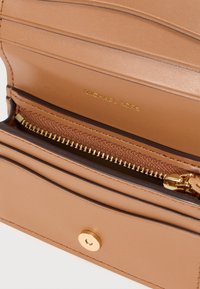 Portefeuille en cuir fauve avec une fermeture éclair dorée, plusieurs emplacements pour cartes, et une fermeture à pression, avec le nom de la marque embossé à l'intérieur.