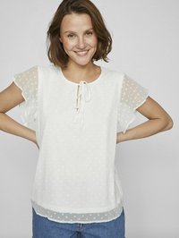 Blusa bianca con motivo a pois, maniche a sbuffo trasparenti, dettagli di collo legato e vestibilità rilassata, abbinata a jeans di denim blu.