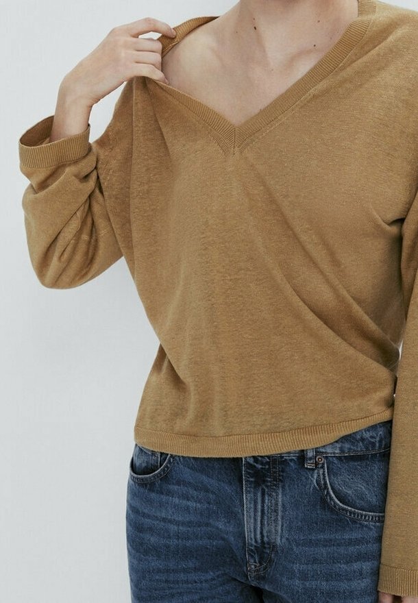 Personne portant un pull col en V marron et un jean bleu, tirant légèrement l'encolure de l'épaule avec la main gauche.