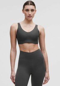 Pelēka sporta krūštura ar dziļu V-veida kakla izgriezumu un platām siksnām, apvienots ar augsto jostasvietu pelēkiem legingiem. Gluda tekstūra bez redzamiem elementiem.