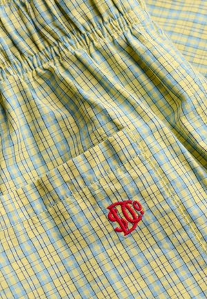 Gult og blåt ternet stof med elastisk talje og lomme med rødt broderet stiliseret logo.