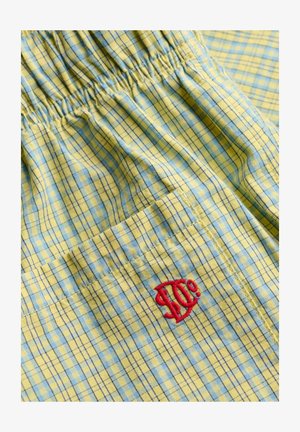 Gult og blåt ternet stof med elastisk talje og lomme med rødt broderet stiliseret logo.