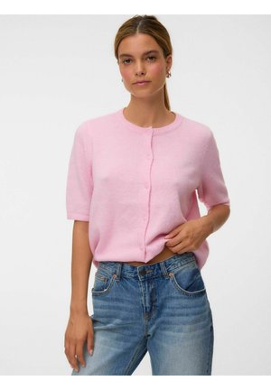 Femme aux cheveux châtain clair attachés, portant un pull rose clair à manches courtes boutonné et un jean bleu, posant devant un fond uni.