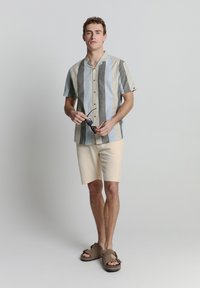 Korte mouwen, gestreept shirt in zachte blauw-, grijs- en beigetinten; gecombineerd met beige shorts en bruine instap sandalen.