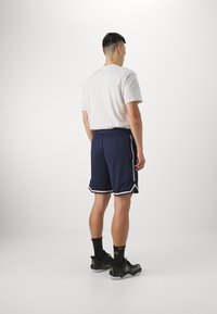 Pantalones cortos deportivos azul marino con franjas laterales blancas, combinados con una camiseta de patrón gris claro y zapatillas negras, vistos desde atrás.