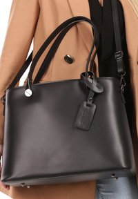 Sac à main en cuir noir avec deux longues anses, forme plate et une étiquette de bagage. Texture lisse avec des accents matériels minimaux.