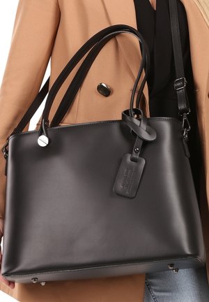 Sac à main en cuir noir avec deux longues anses, forme plate et une étiquette de bagage. Texture lisse avec des accents matériels minimaux.