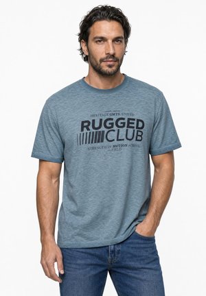 Homme aux cheveux courts et foncés avec une barbe, portant un t-shirt bleu avec l'inscription "Rugged Club" et un jean bleu, debout avec une main dans la poche.